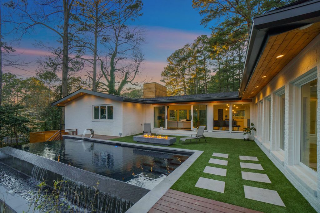 A luxurious infinity pool with a fire pit, set beside a modern home with large windows, viewed during sunset.