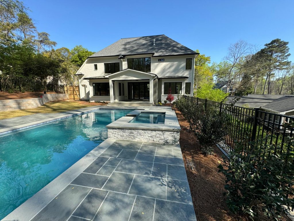 A newly built pool with stone decking, surrounded by a spacious yard and a modern white house in Atlanta.