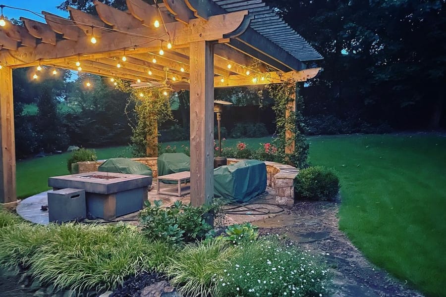 A cozy outdoor patio at dusk, featuring a wooden pergola adorned with string lights. The space includes a covered seating area around a stone fire pit, surrounded by lush greenery.