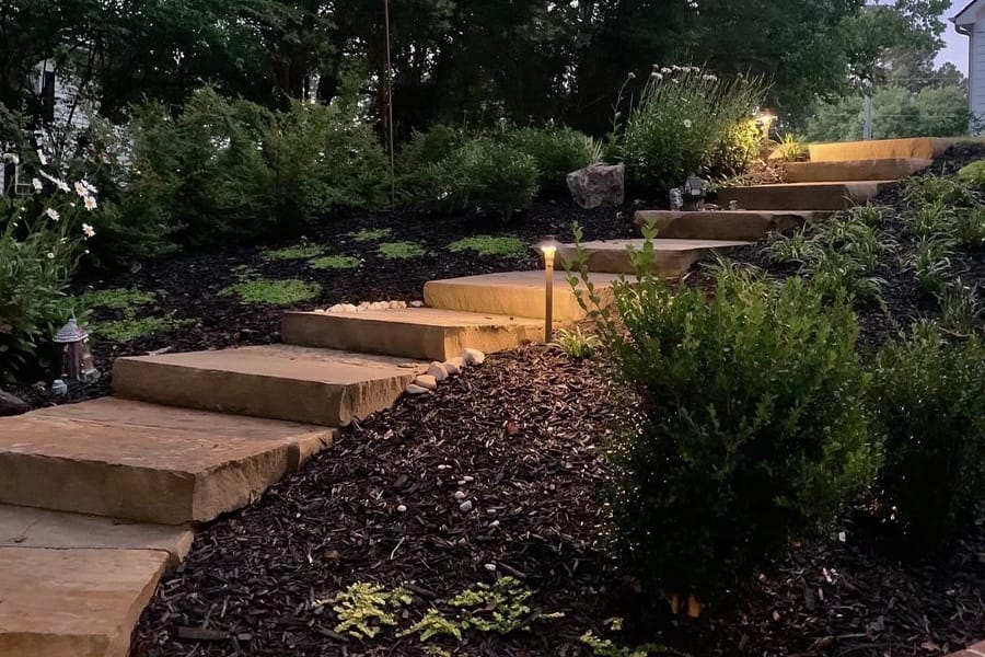 Stone steps in a landscaped garden, softly lit by pathway lights at dusk. The scene is serene, surrounded by lush greenery and small bushes.