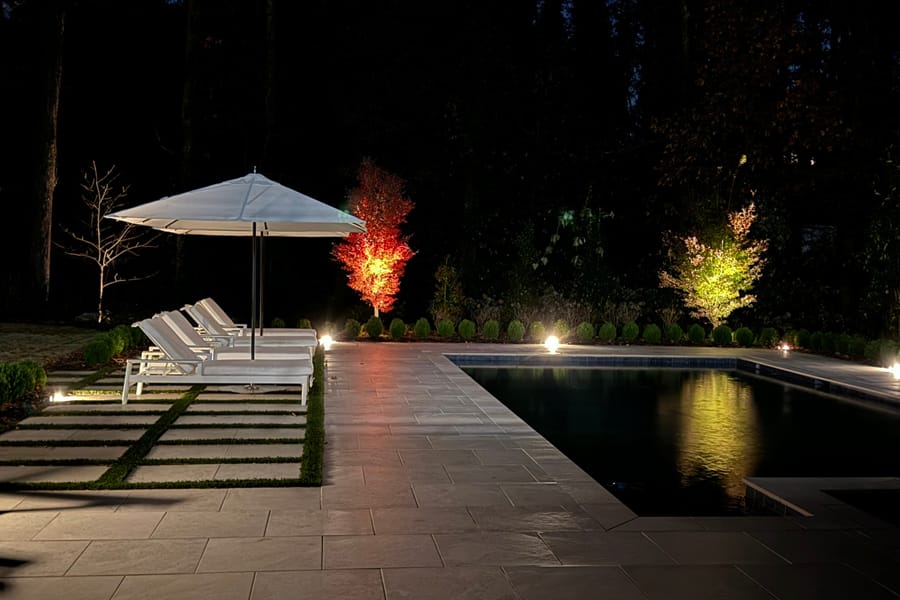 Nighttime poolside scene with two white lounge chairs under an umbrella, lit by soft spotlights. Trees glow with red and yellow lights, reflecting a serene ambience.