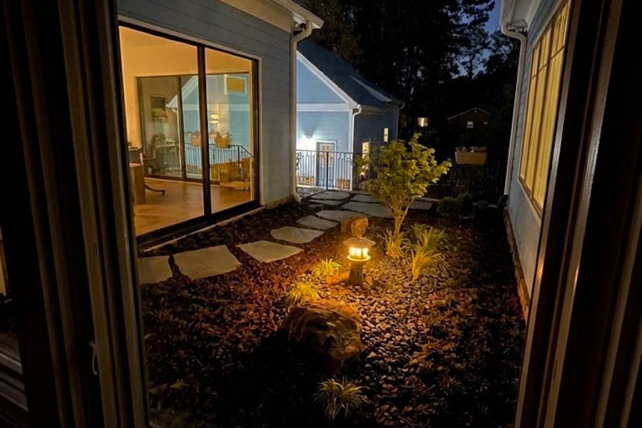Night view of a cozy patio garden with stepping stones and a glowing lamp. Warm indoor light spills from large windows, creating a serene atmosphere.