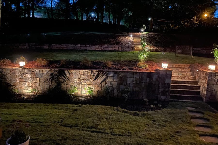 Tiered garden at night with stone walls, illuminated by glowing lanterns on each level. Steps lead up through lush greenery, creating a serene ambiance.