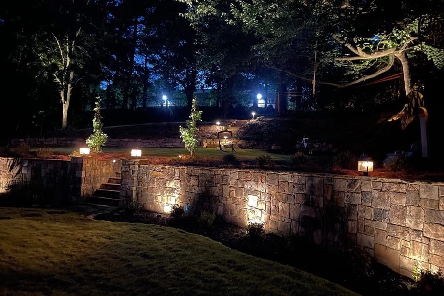 A serene, dimly lit garden at night, featuring illuminated stone retaining walls, trees, and lanterns. The scene conveys a peaceful ambiance.